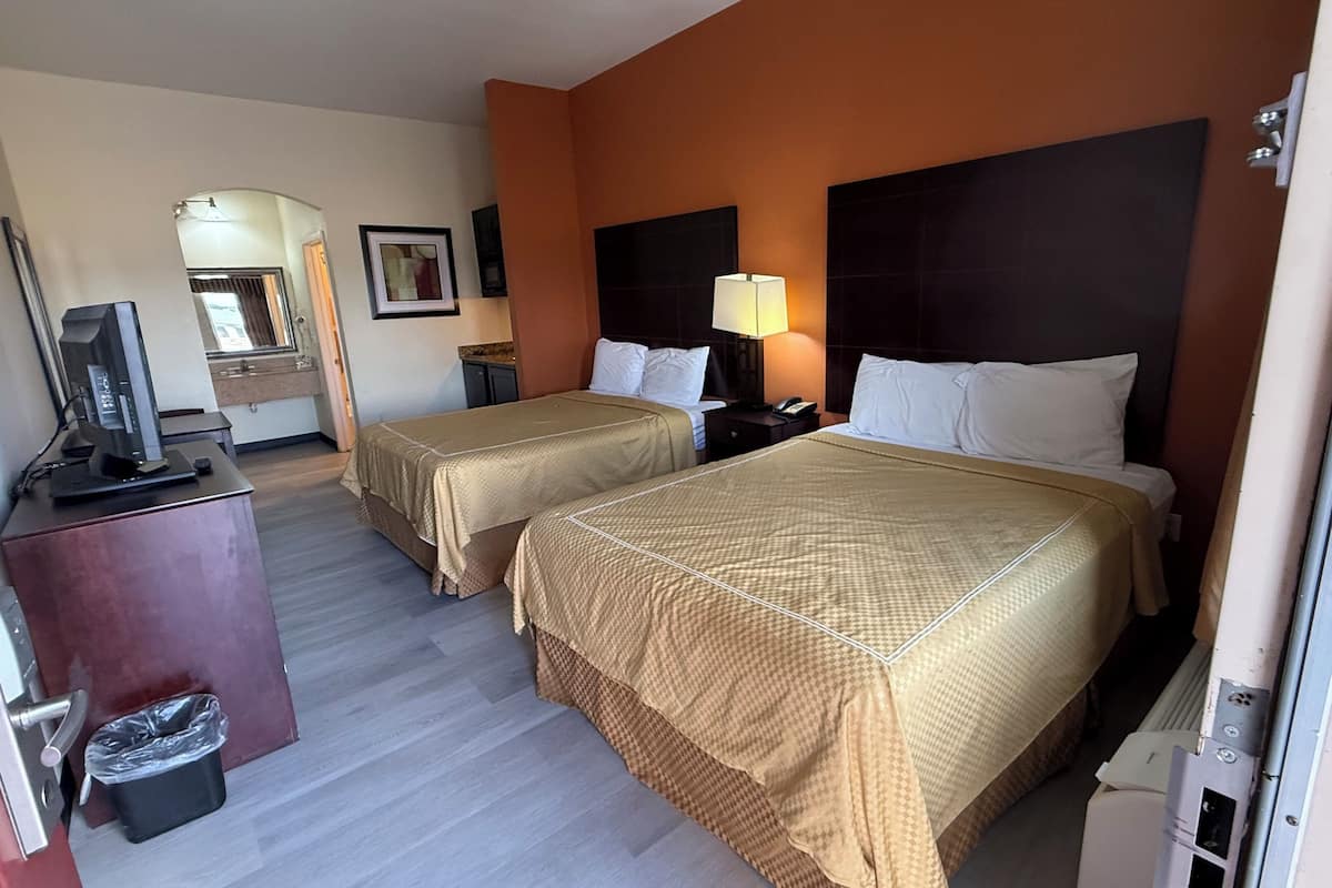 A hotel room with two beds, a desk with a computer, a trash bin, and a bathroom visible through an archway.