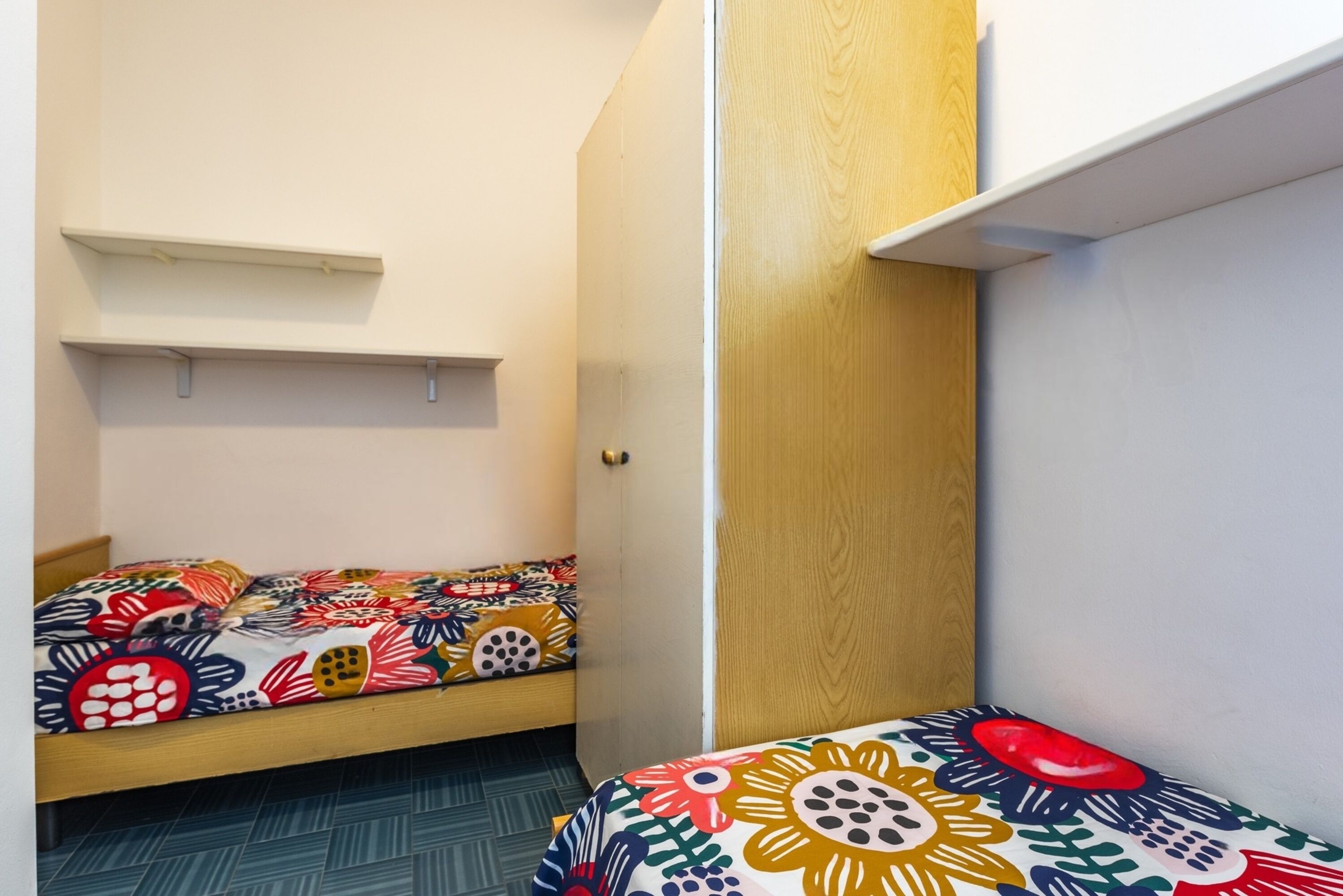 Une chambre de dortoir avec deux lits superposés, chacun agrémenté d’une literie aux motifs floraux colorés. Des étagères en bois se trouvent au-dessus des lits, et une porte est située sur le côté droit.