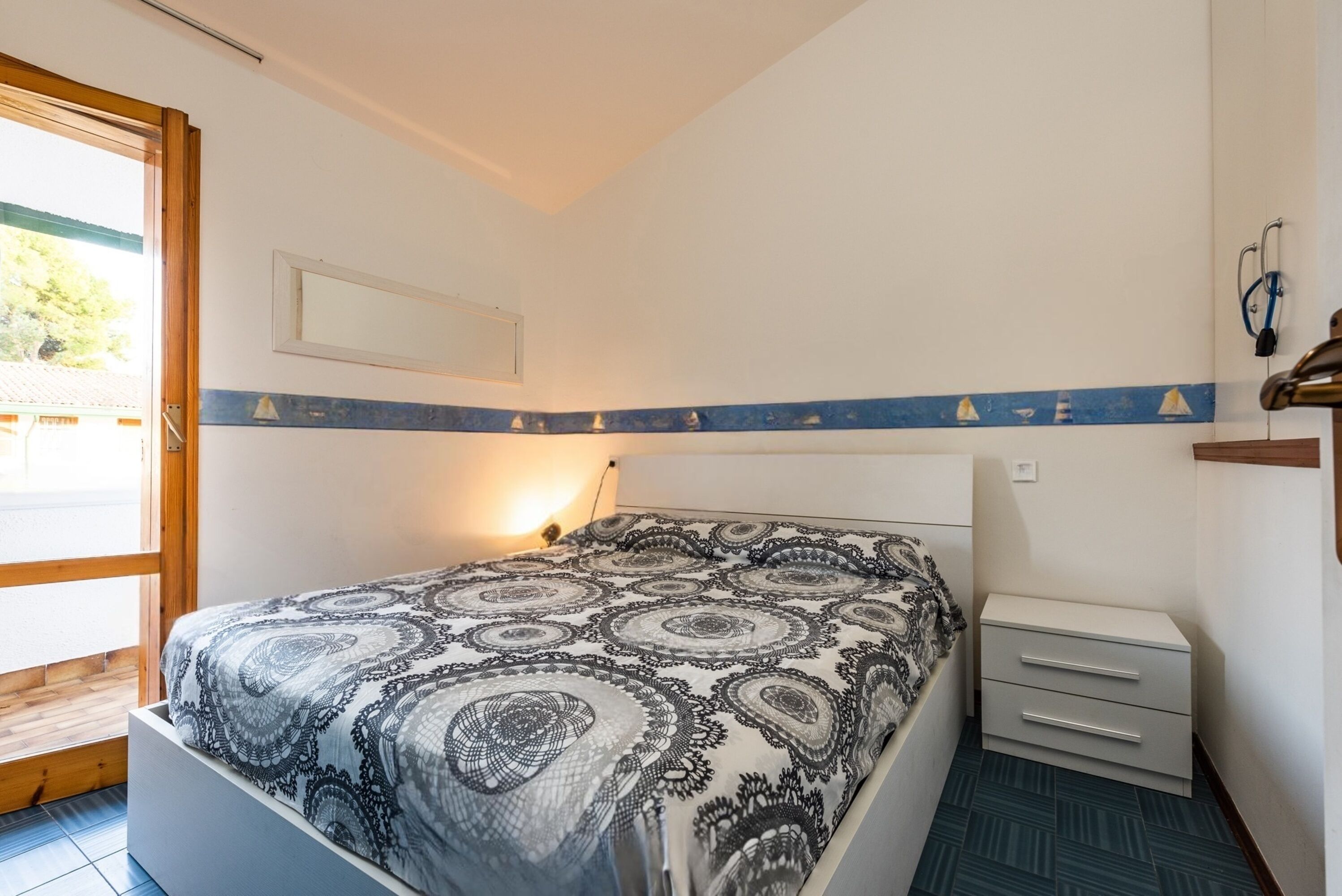 Ein Schlafzimmer mit gemustertem Bettbezug, einem Nachttisch mit Schubladen, einem an der Wand befestigten Regal und einem Fenster mit Aussicht.