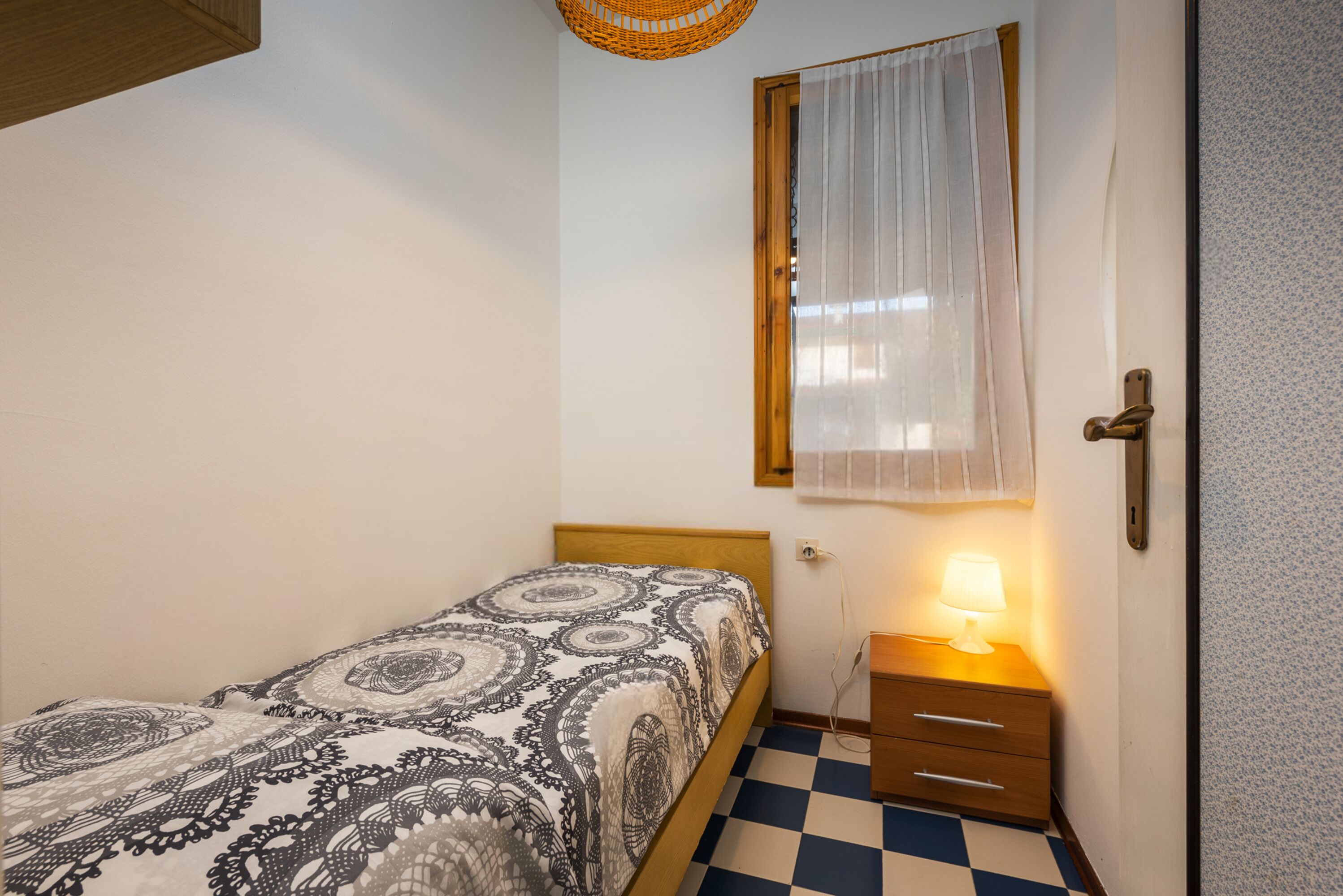 Un dormitorio pequeño con piso a cuadros, una cama con colcha estampada, una mesita de noche con lámpara y una ventana con cortina translúcida.