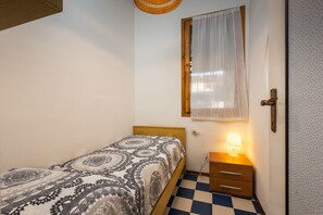 Un dormitorio pequeño con piso a cuadros, una cama con colcha estampada, una mesita de noche con lámpara y una ventana con cortina translúcida.