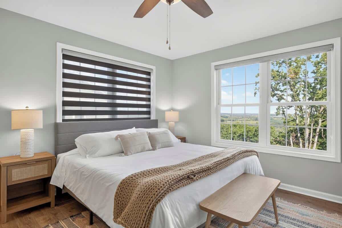 A bedroom with a bed, bedside lamps, a ceiling fan, a window with a view, and a bench.