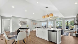 A modern kitchen with a central island, white cabinetry, and a dining area with a round table and chairs. - Mountain View Cottage | Hot Tub & Game Room (Front Royal)