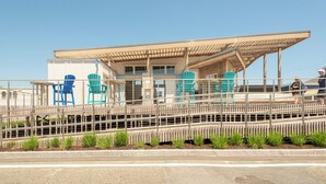 Un edificio frente a la playa con un área de estar techada, varias sillas azules y una valla de madera.
