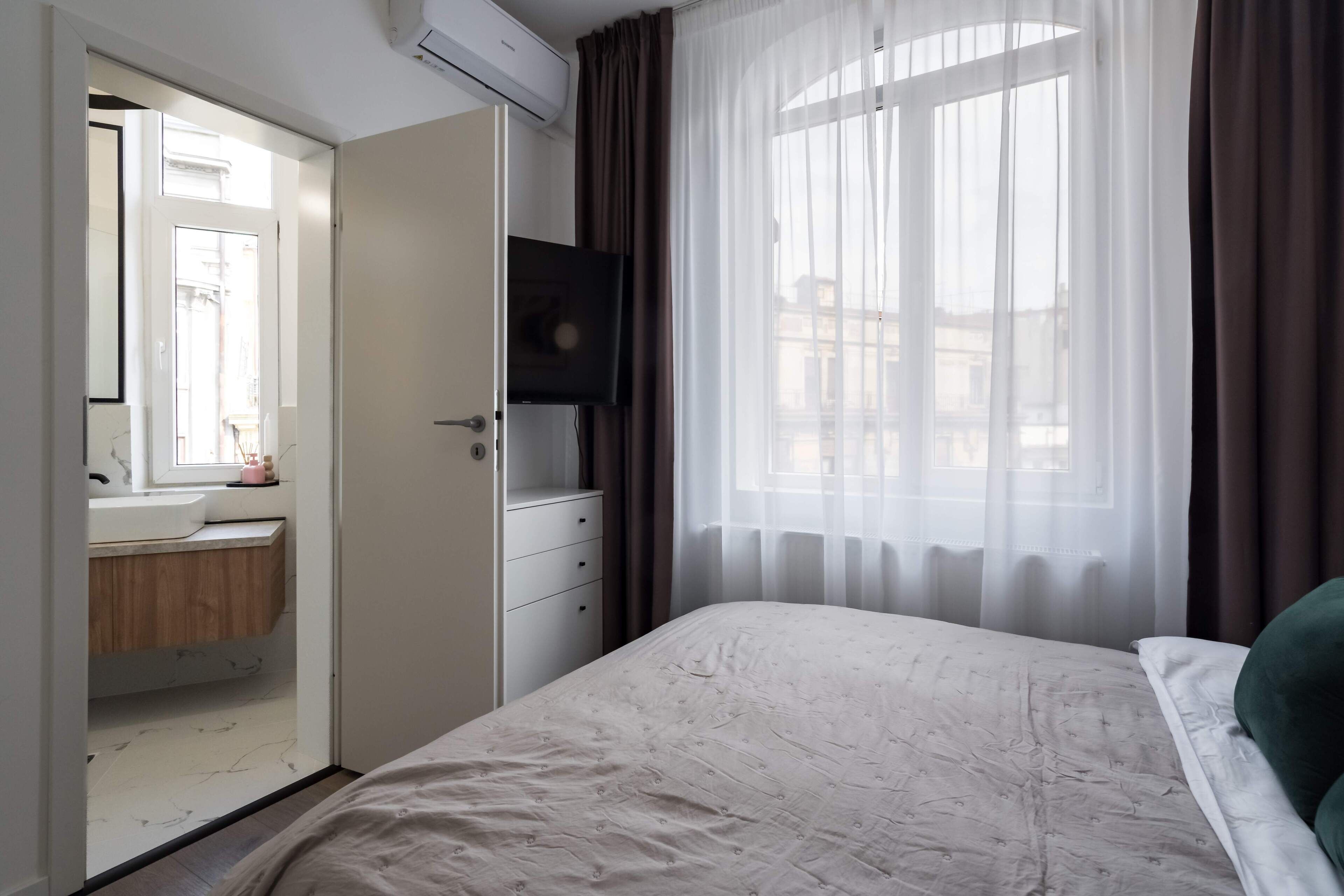 A bedroom with a bed, a TV mounted on the wall, a window with sheer curtains, and a bathroom visible through an open door.