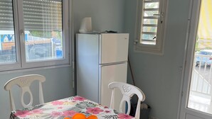 A dining area with a table covered in a floral tablecloth, white chairs with blue cushions, a refrigerator, and windows with blinds. - Alamanda pink (Sainte-Anne)