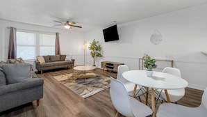 Una sala moderna con un sofá gris, una mesa de centro redonda y una zona de comedor con sillas y una mesa blancos.