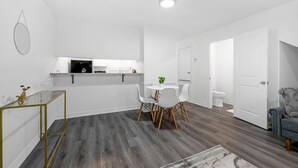 Una cocina moderna con gabinetes blancos, un comedor con una mesa redonda y sillas, y un sofá gris en una habitación luminosa de paredes blancas y suelo de madera.