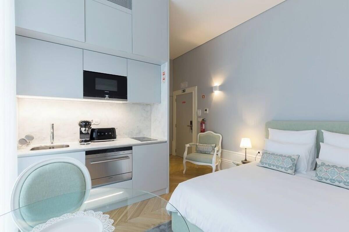 A compact hotel room with a kitchenette, a bed with matching pillows, a chair, and a bedside table with a lamp.