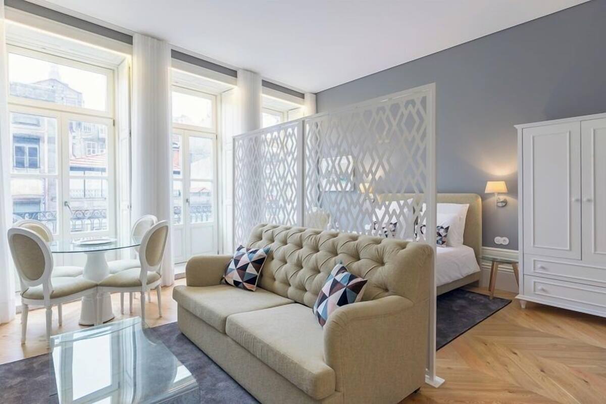 A modern bedroom with a large window, a tufted sofa, a glass coffee table, and a white wardrobe.