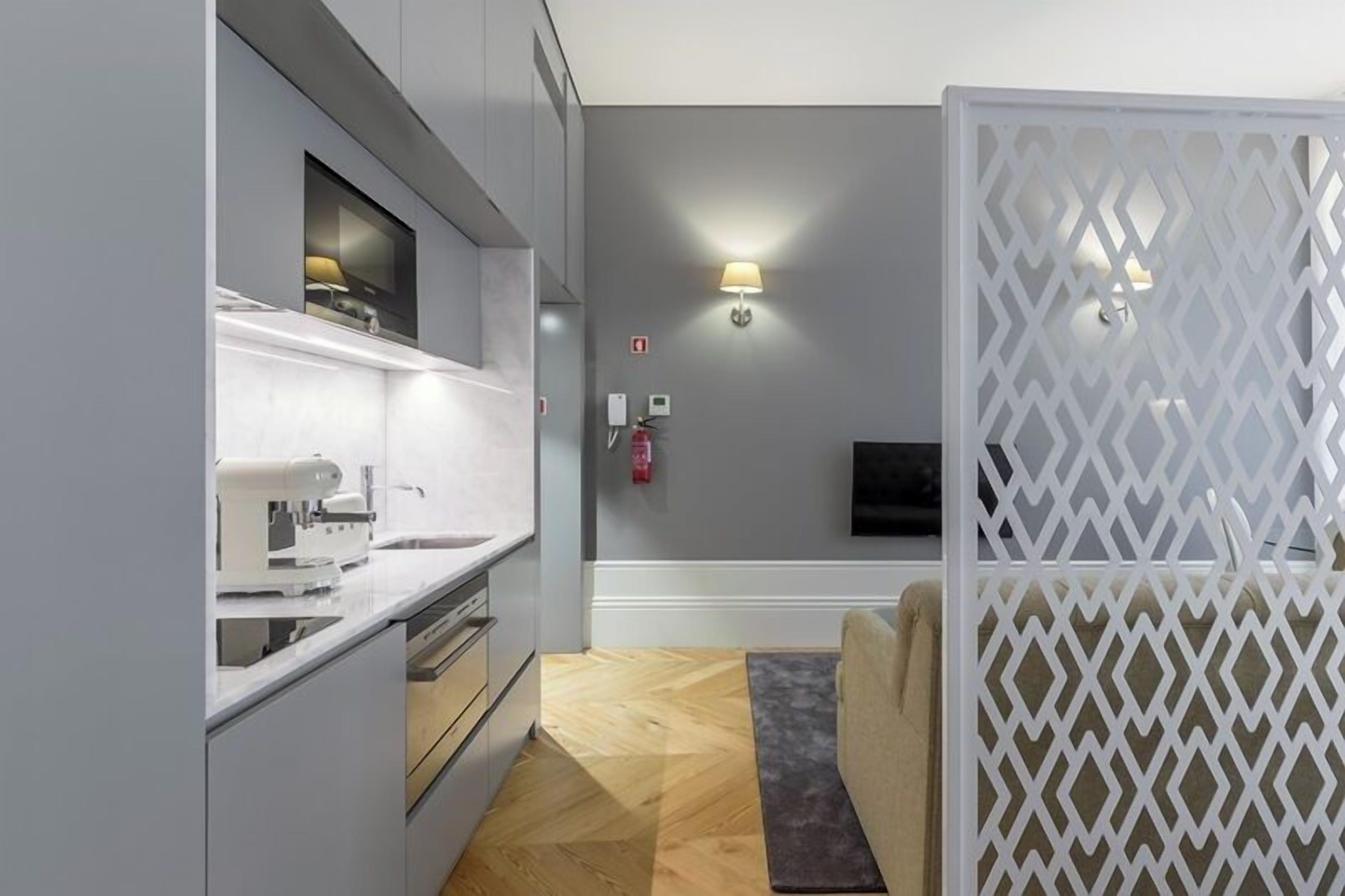 A modern kitchen with a built-in oven, a microwave, and a sink. There is a white partition with a geometric pattern, a wall-mounted TV, and a wall-mounted light fixture.
