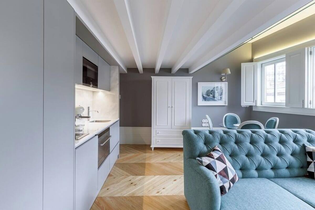 A modern kitchen with white cabinets, a herringbone wood floor, and a teal sofa with patterned cushions.