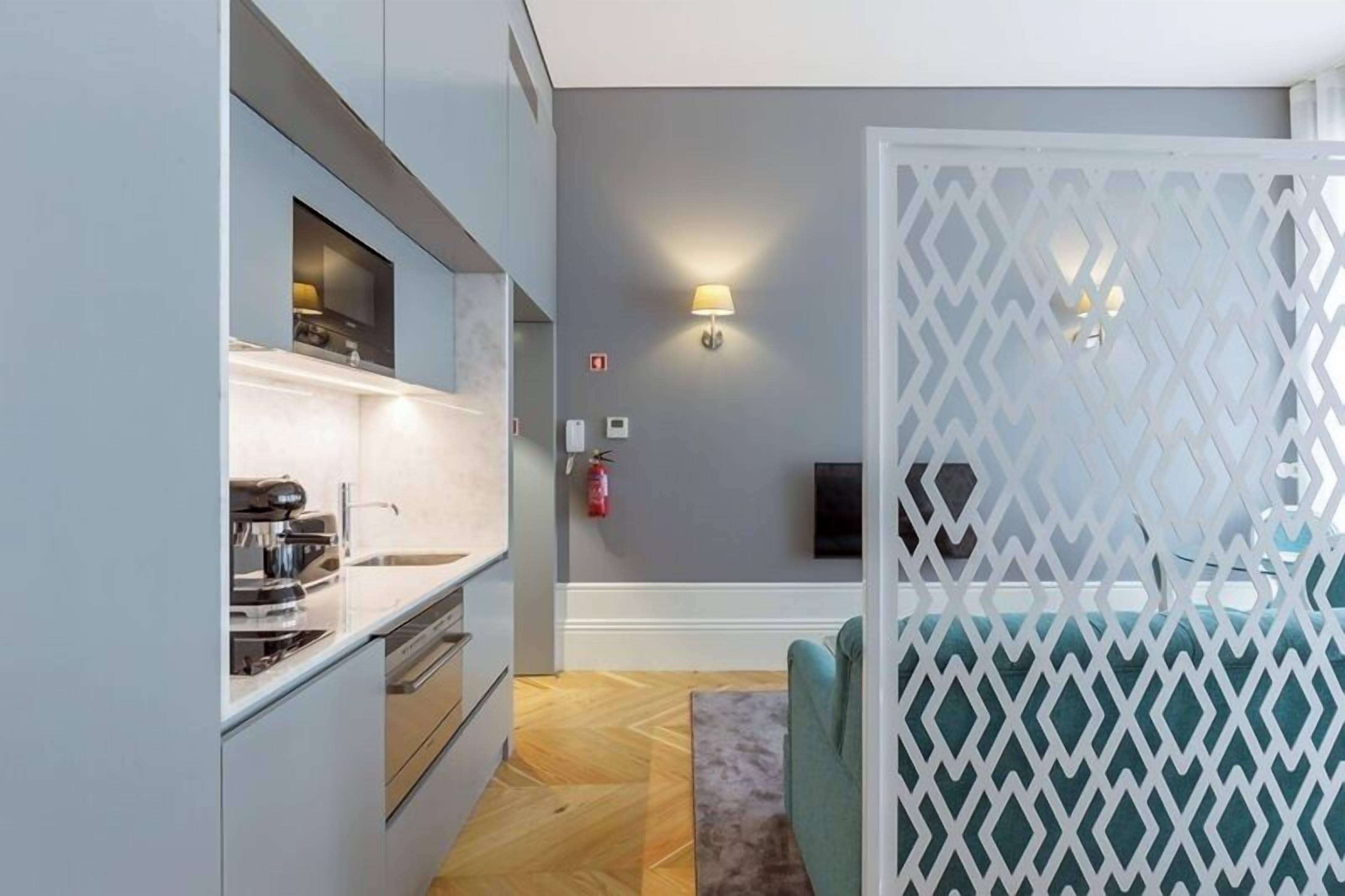 A modern kitchen with built-in appliances, a dining area separated by a white partition, and a grey wall with a wall-mounted lamp.