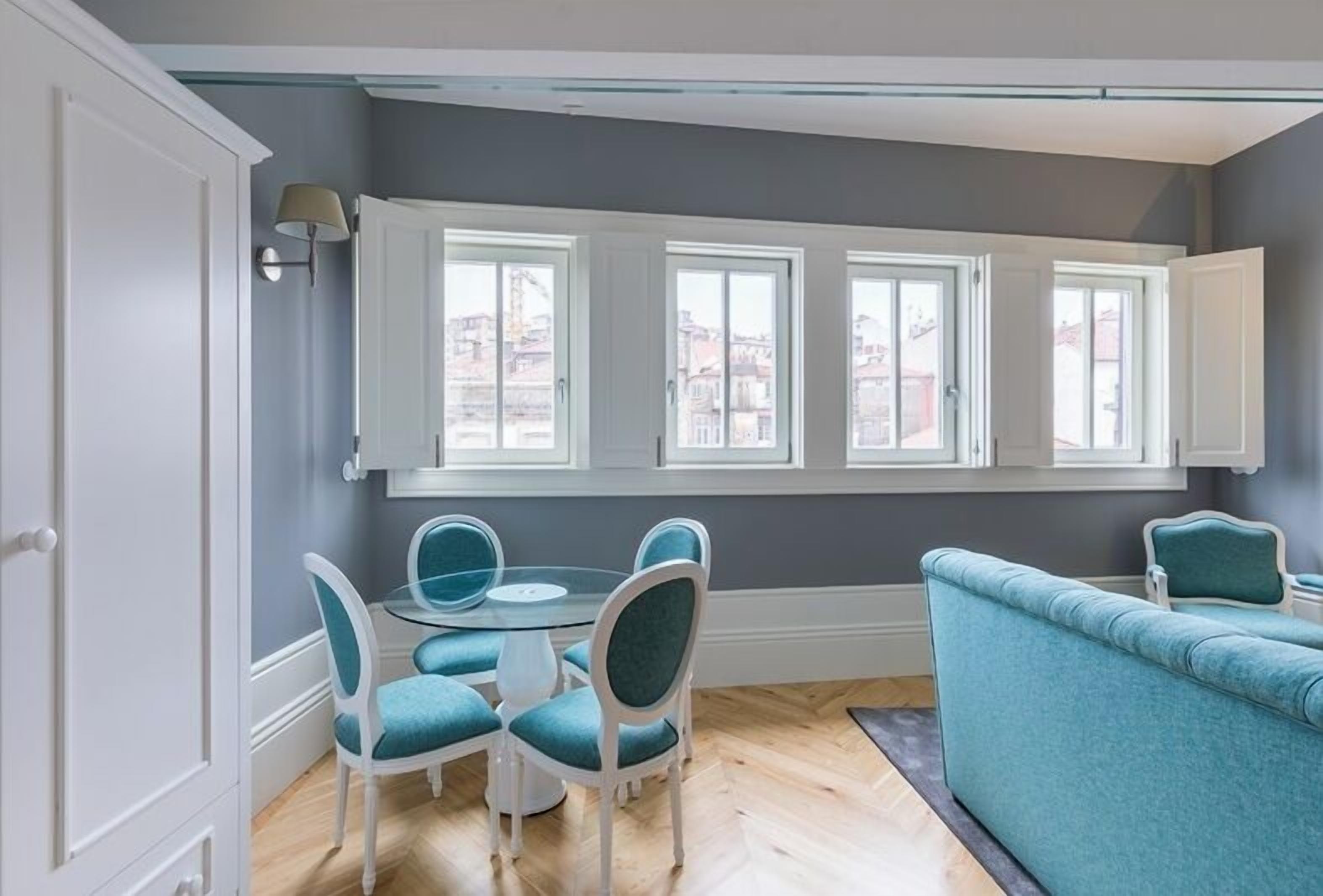 A dining area with a round table and four chairs, a sofa, and large windows with white frames.