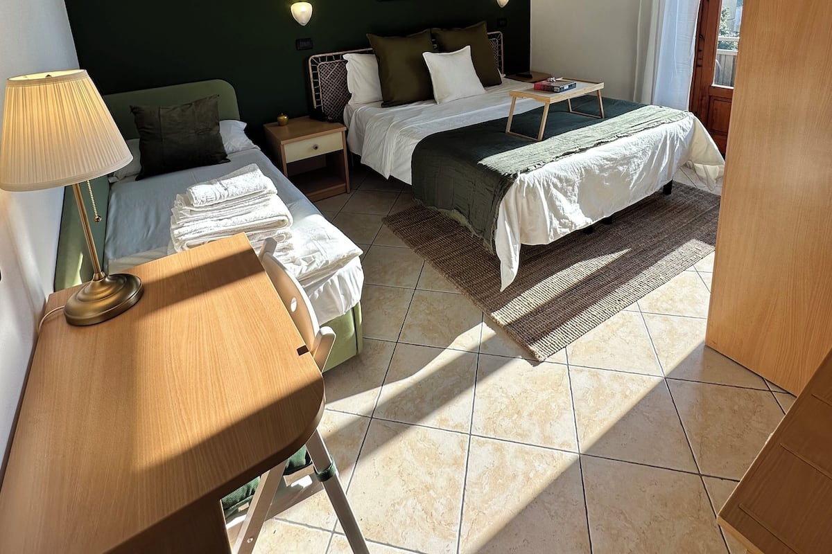 Ein Hotelzimmer mit zwei Betten, einem Schreibtisch mit Lampe, einem Nachttisch, einem kleinen Tisch mit einer Vase und einem Fenster mit Vorhängen.