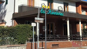 Ein zweistöckiges Gebäude mit einem Schild, auf dem „La Riviera“ steht. Es verfügt über Balkone, einen überdachten Außenbereich und Schilder, die auf nahegelegene Sehenswürdigkeiten wie Miramonti und Camping Belsito hinweisen.