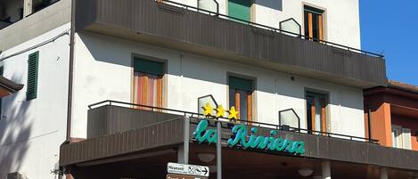 Un edificio de dos pisos con un cartel que dice "La Riviera". Tiene balcones, una zona de estar al aire libre techada, y carteles que señalan atracciones cercanas como Miramonti y Camping Belsito.