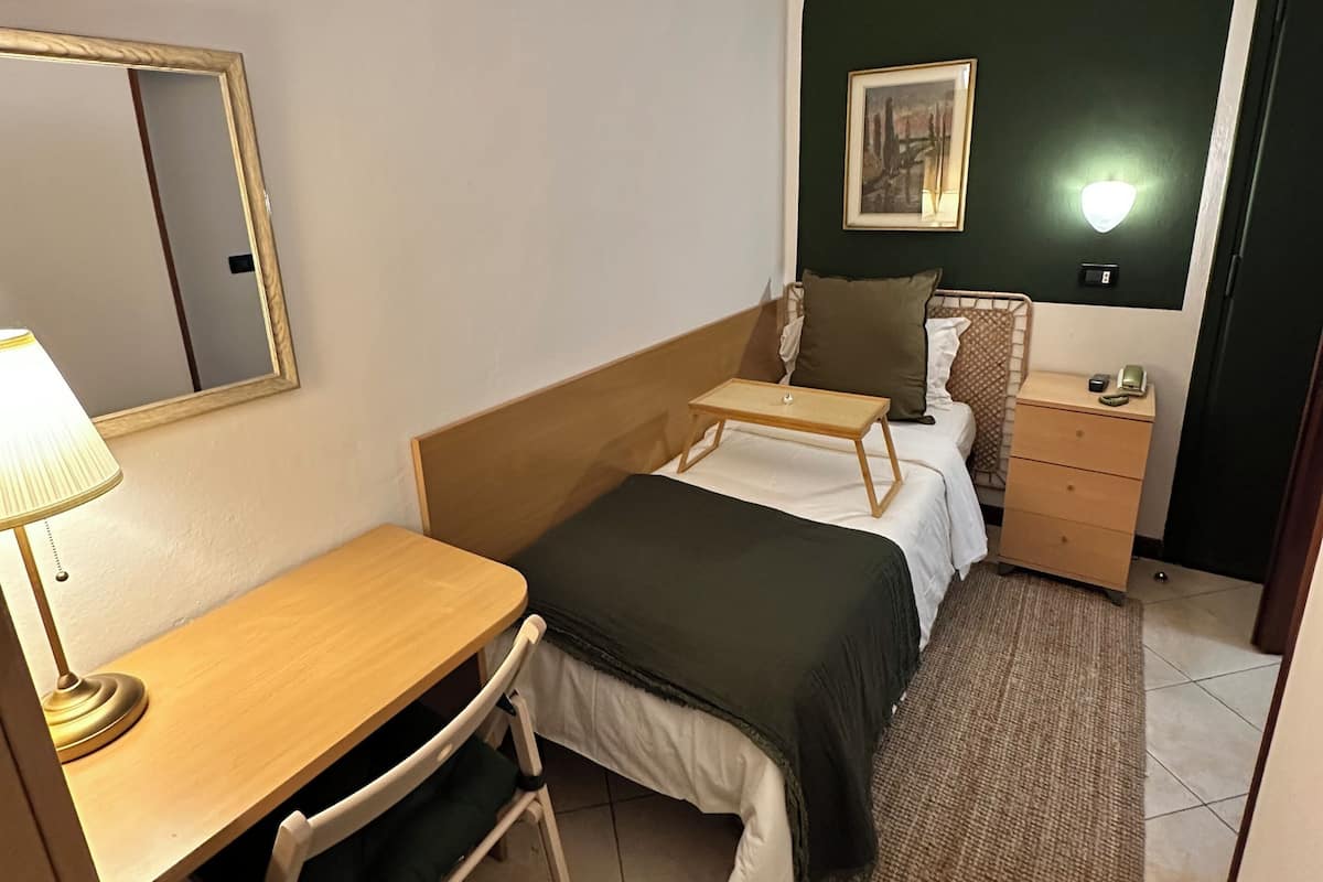 Ein kleines Hotelzimmer mit Bett, Nachttisch, Lampe, Stuhl und einem kleinen Schreibtisch.