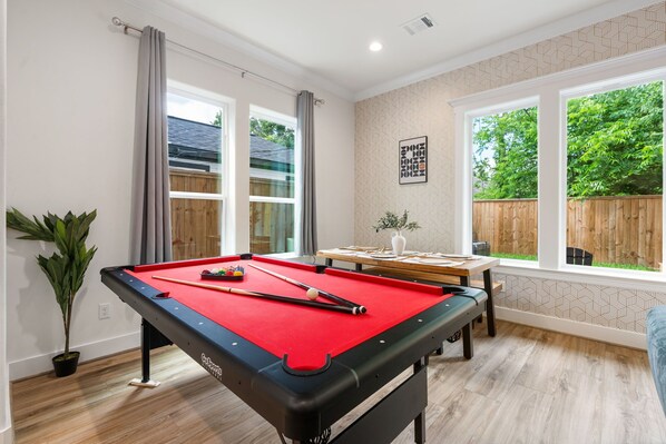 Une table de billard recouverte d’un feutre rouge, une table en bois ornée d’un vase et une fenêtre donnant sur une clôture en bois et de la verdure.