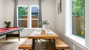 Een eetgedeelte met een houten tafel voor zes personen, een pooltafel en een raam met uitzicht op een achtertuin.