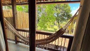 A hammock on a wooden deck with a view of a green garden and a yellow building. - Pousada Vilarejo (Itacaré)