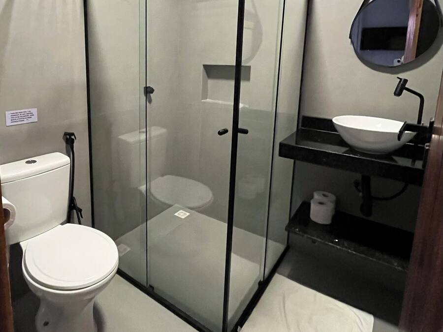 Un bany modern amb dutxa d'acer, un vàter blanc i un lavabo negre amb un mirall rodó a dalt.