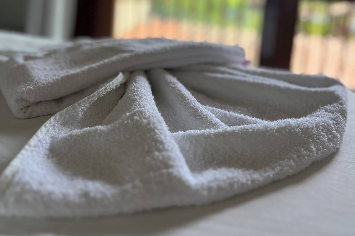 A neatly folded white towel on a bed with a view of greenery outside through a window.