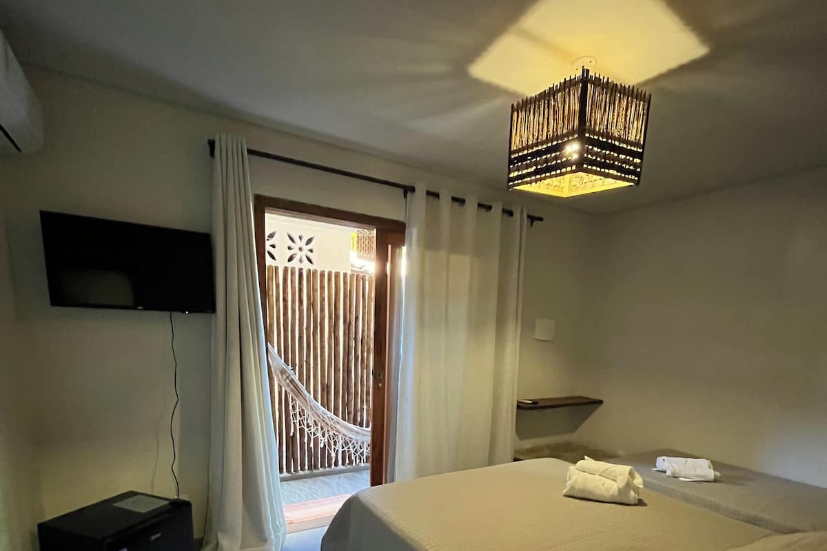 A hotel room with a bed, a TV mounted on the wall, a desk with a lamp, and a view of the outside through a window with curtains.