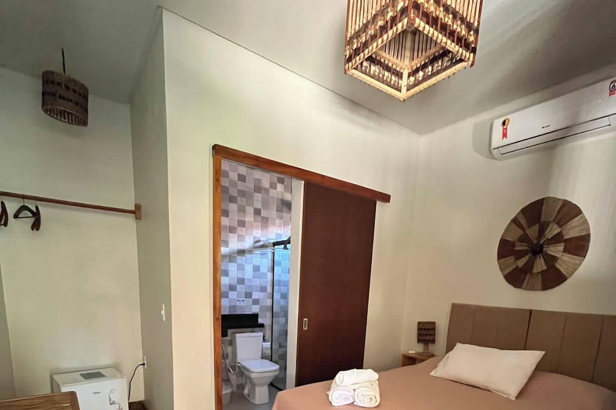 A bedroom with a bed, a wooden headboard, a bedside table, a wall-mounted air conditioner, and a door leading to a bathroom.