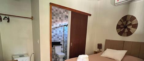 A bedroom with a bed, a wooden headboard, a bedside table, a wall-mounted air conditioner, and a door leading to a bathroom.