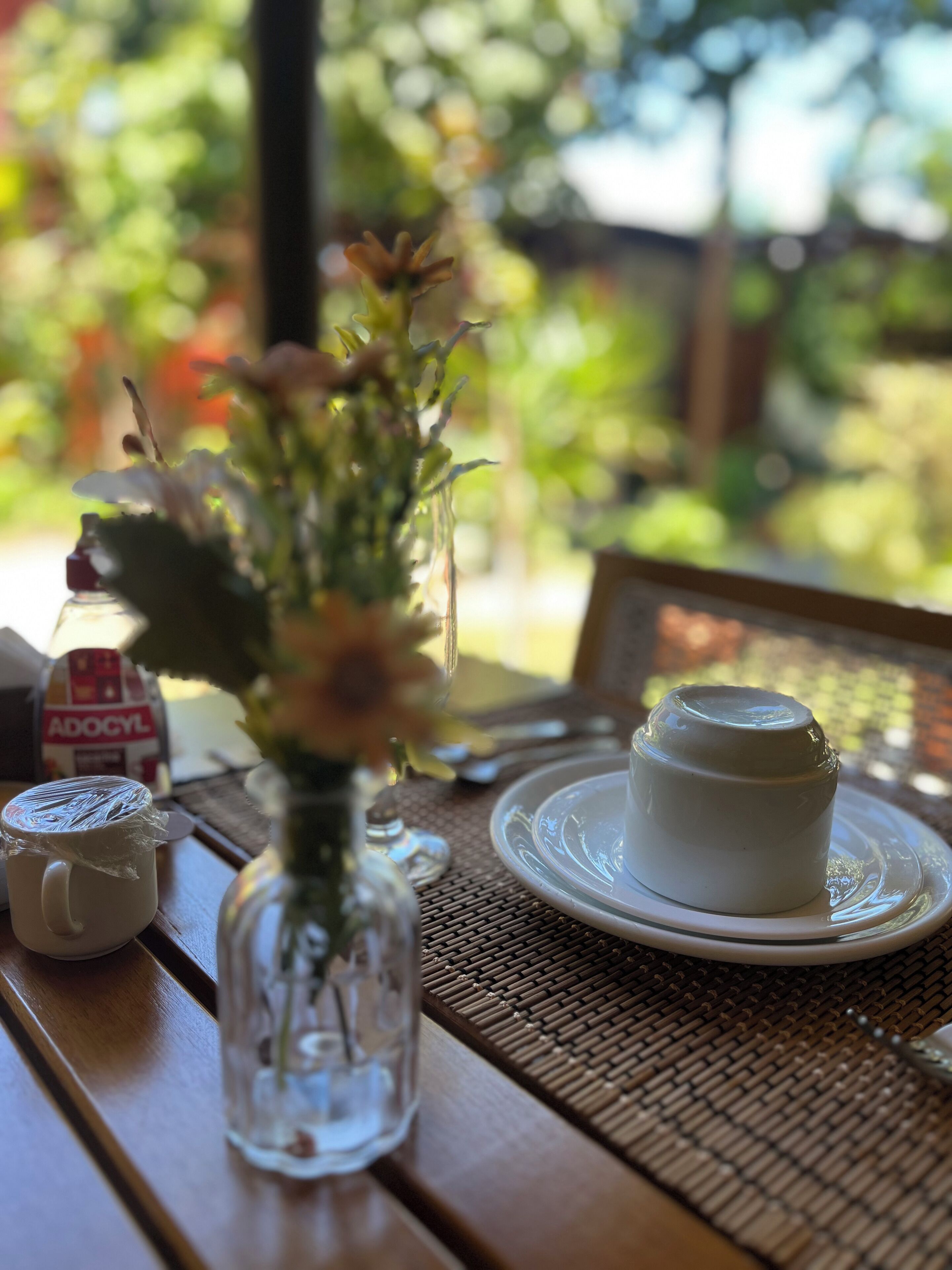 Una mesa con un jarrón de vidrio transparente con flores, una taza blanca con film plástico, una taza de porcelana blanca y un plato con una taza blanca encima.