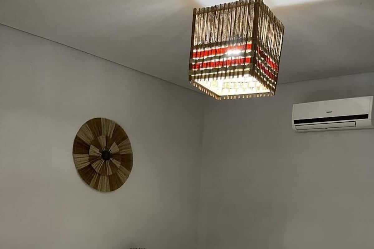 A hotel room with two beds, beige bedding, white pillows, a decorative wall hanging, a hanging light fixture, and an air conditioning unit.
