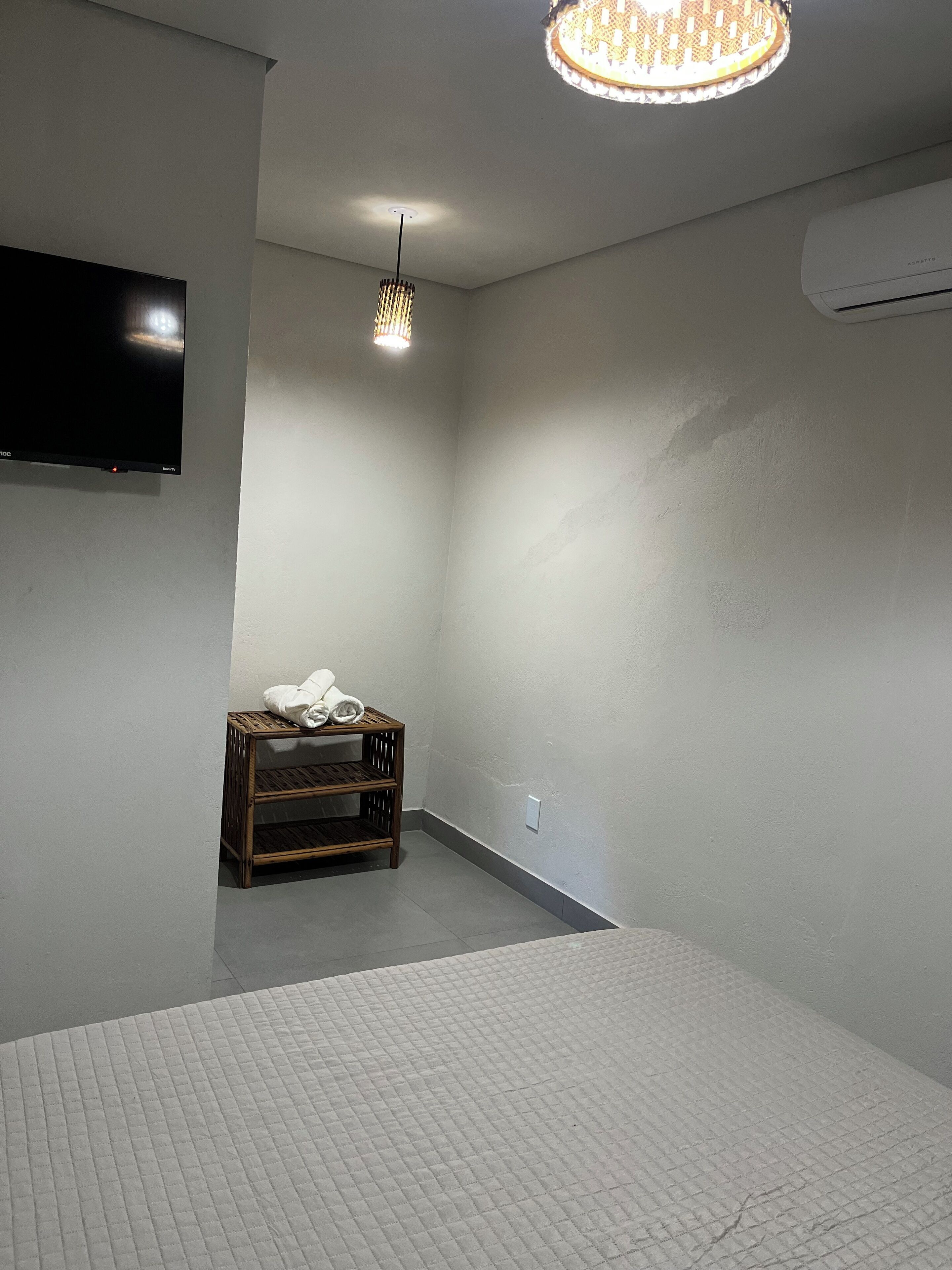 Una habitación con una cama, un televisor montado en la pared, una lámpara colgante y un pequeño estante de madera con toallas.