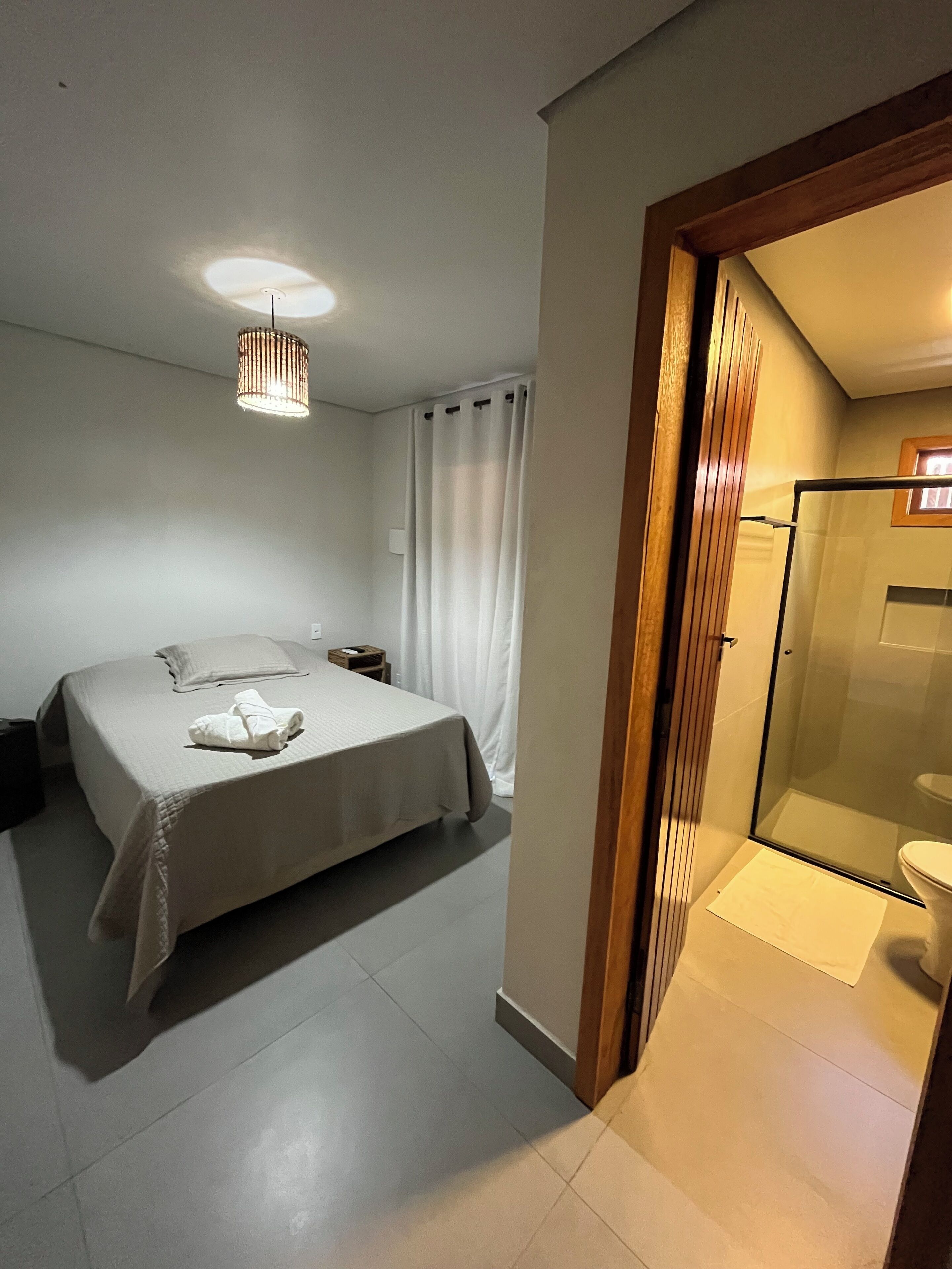 A hotel room with a bed, a bedside table, a nightstand, a hanging light fixture, a shower area with a glass door, and a toilet.