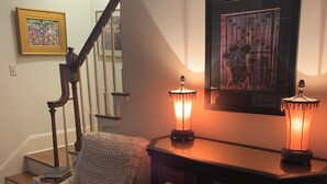 Uma sala de estar aconchegante com uma poltrona branca, uma mesa de madeira com duas luminárias, uma obra de arte emoldurada na parede e uma escada ao fundo.