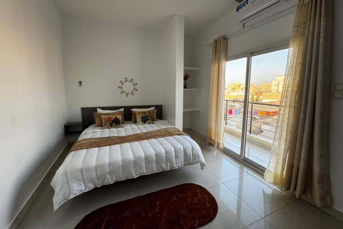 A modern bedroom with a large bed, a dark headboard, a small shelf unit, and a view of the city through a large window with curtains.