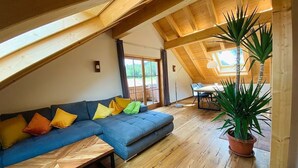 Een gezellige loft-achtige kamer met een houten plafond, een blauwe modulaire bank, een houten salontafel en een potplant.