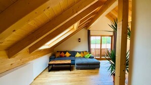 Een loft-achtige kamer met een schuine kap, houten balken, een bank, een salontafel en een potplant.