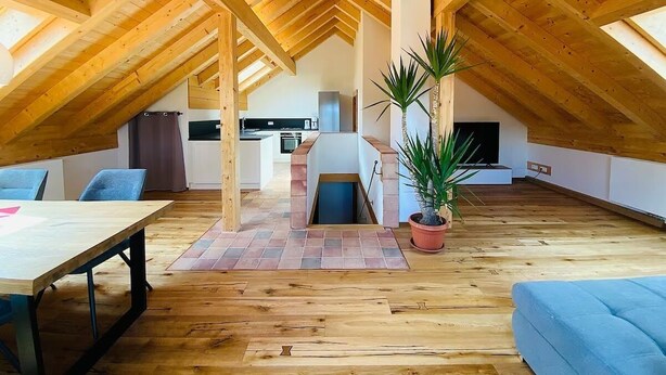 Een ruime zolderkamer met houten balken, een eettafel, blauwe stoelen, een plant in pot en een keuken op de achtergrond.