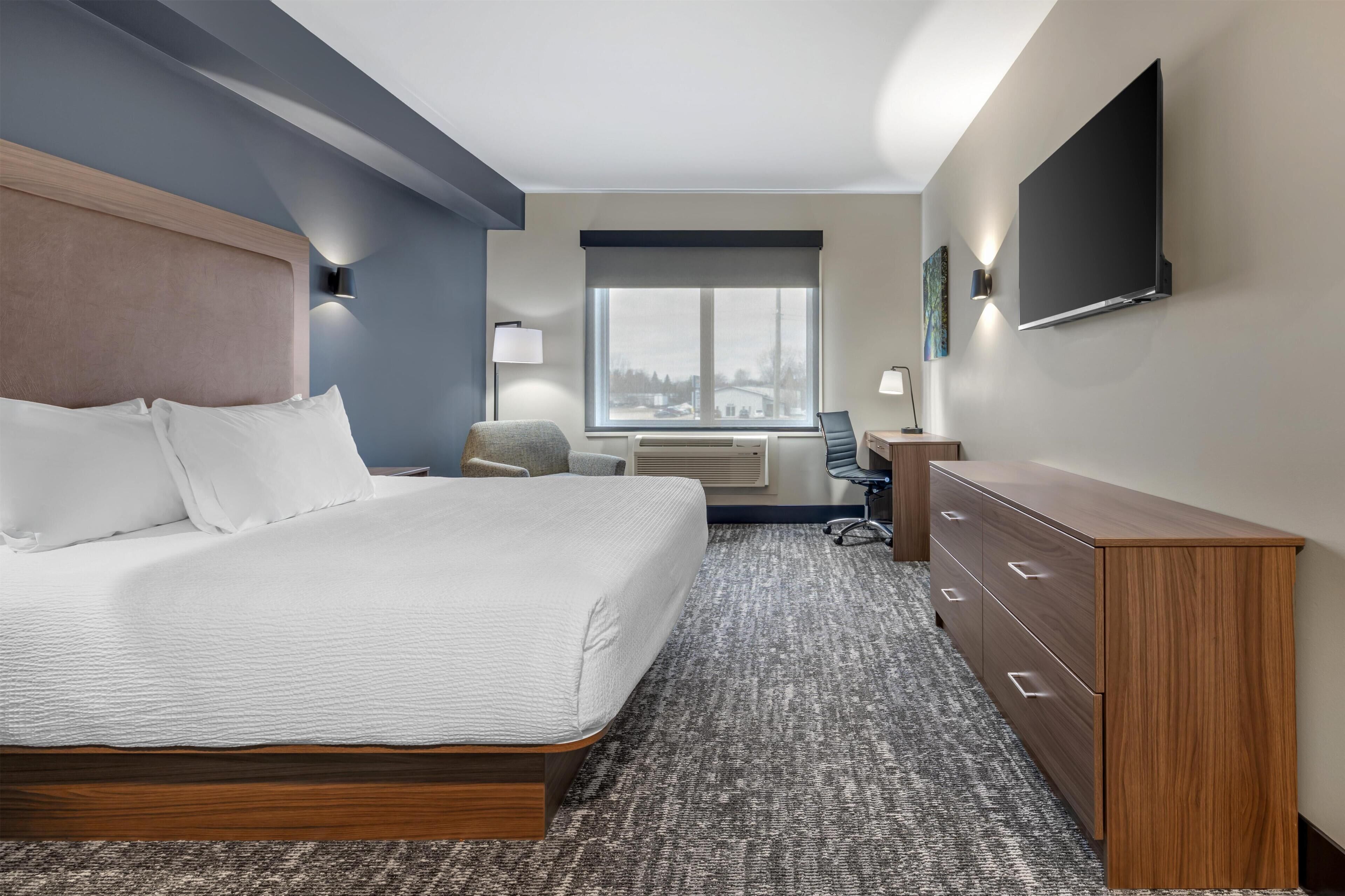 A hotel room with a bed, a desk, a chair, a dresser, a TV mounted on the wall, and a window with a view.