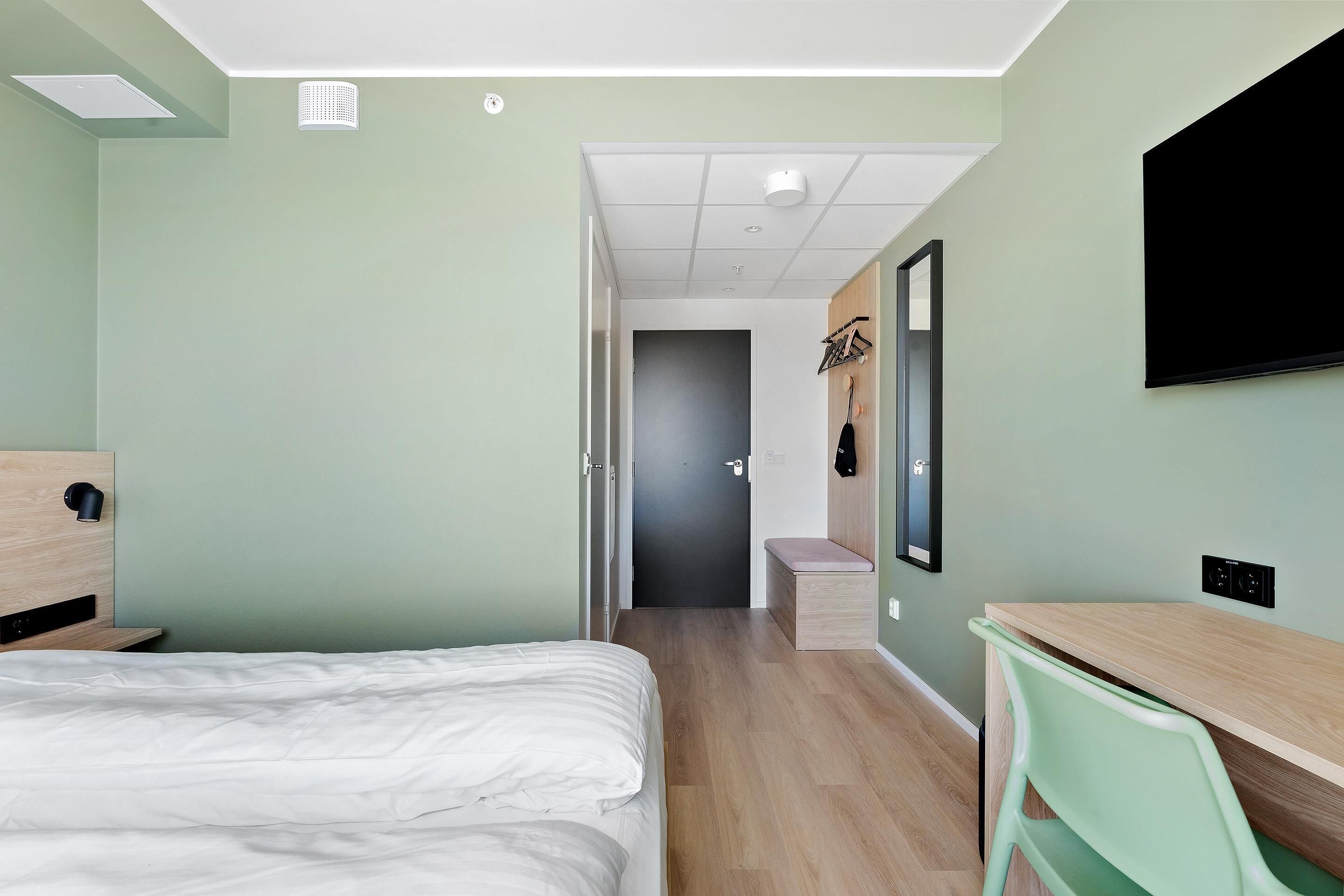 A hotel room with a bed, a desk, a chair, a TV, and a door leading to a bathroom.