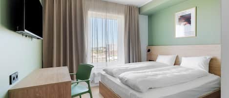 Une chambre d’hôtel avec un lit, un bureau, une chaise, une télévision et une fenêtre avec des rideaux.