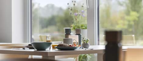 A breakfast table with a bowl, mug, and plate of food, a small plant, and a window with a view of trees and flowers.