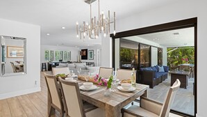 A modern dining area with a wooden table set for four, surrounded by chairs. A chandelier hangs above, and the room opens to a living area with a sofa and coffee table. - Boynton Beach Retreat, Close to Delray, Aug Deal (Boynton Beach)