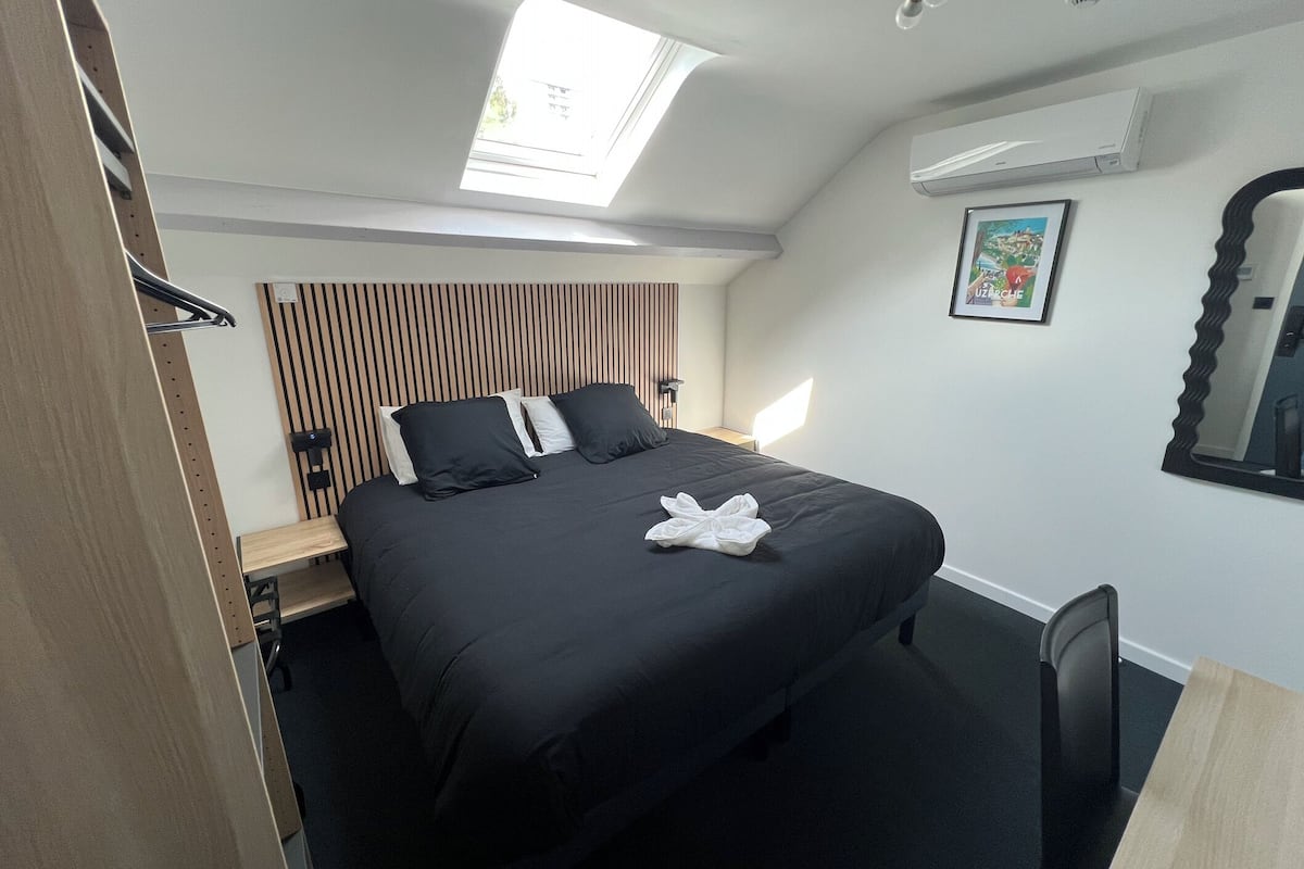 A modern hotel room with a skylight, a bed with black bedding, a bedside table, a chair, and a wall-mounted air conditioner.