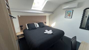 Una habitación de hotel moderna con tragaluz, una cama con ropa de cama negra, una mesita de noche, una silla y un aire acondicionado fijado a la pared.