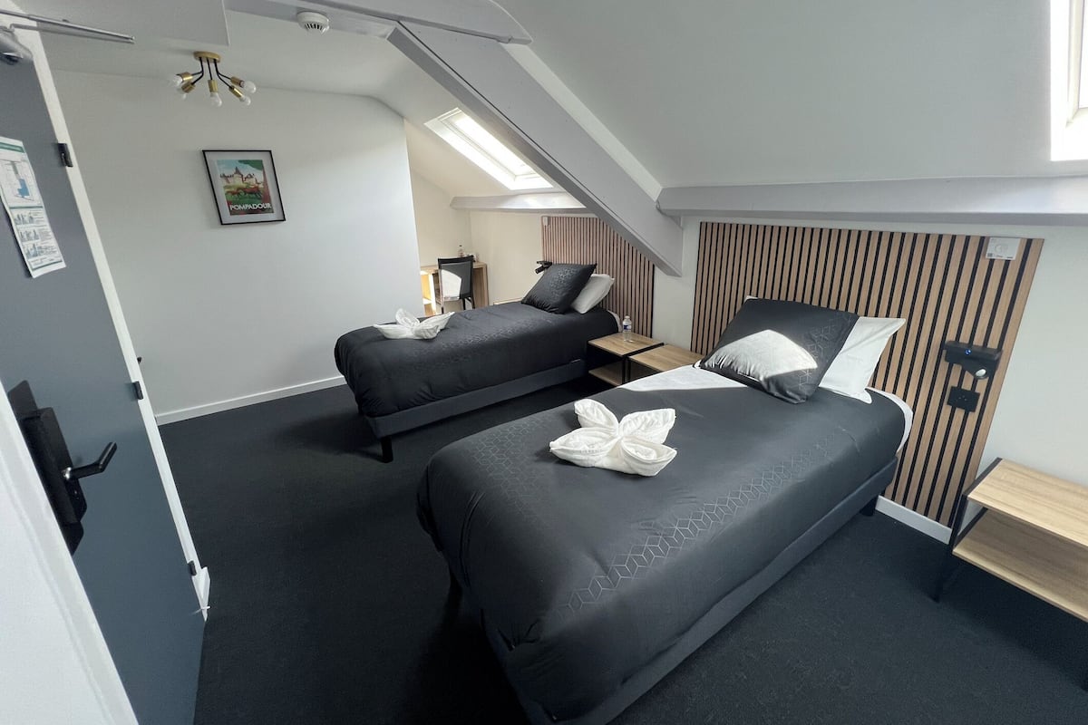 A modern hotel room with two beds, a wooden slat headboard, a small bedside table, and a ceiling light fixture.