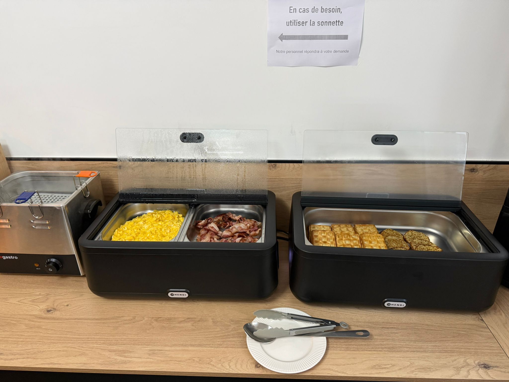 Una estación de buffet con dos dispensadores de comida abiertos, uno con huevos revueltos y bacon, y el otro con gofres.