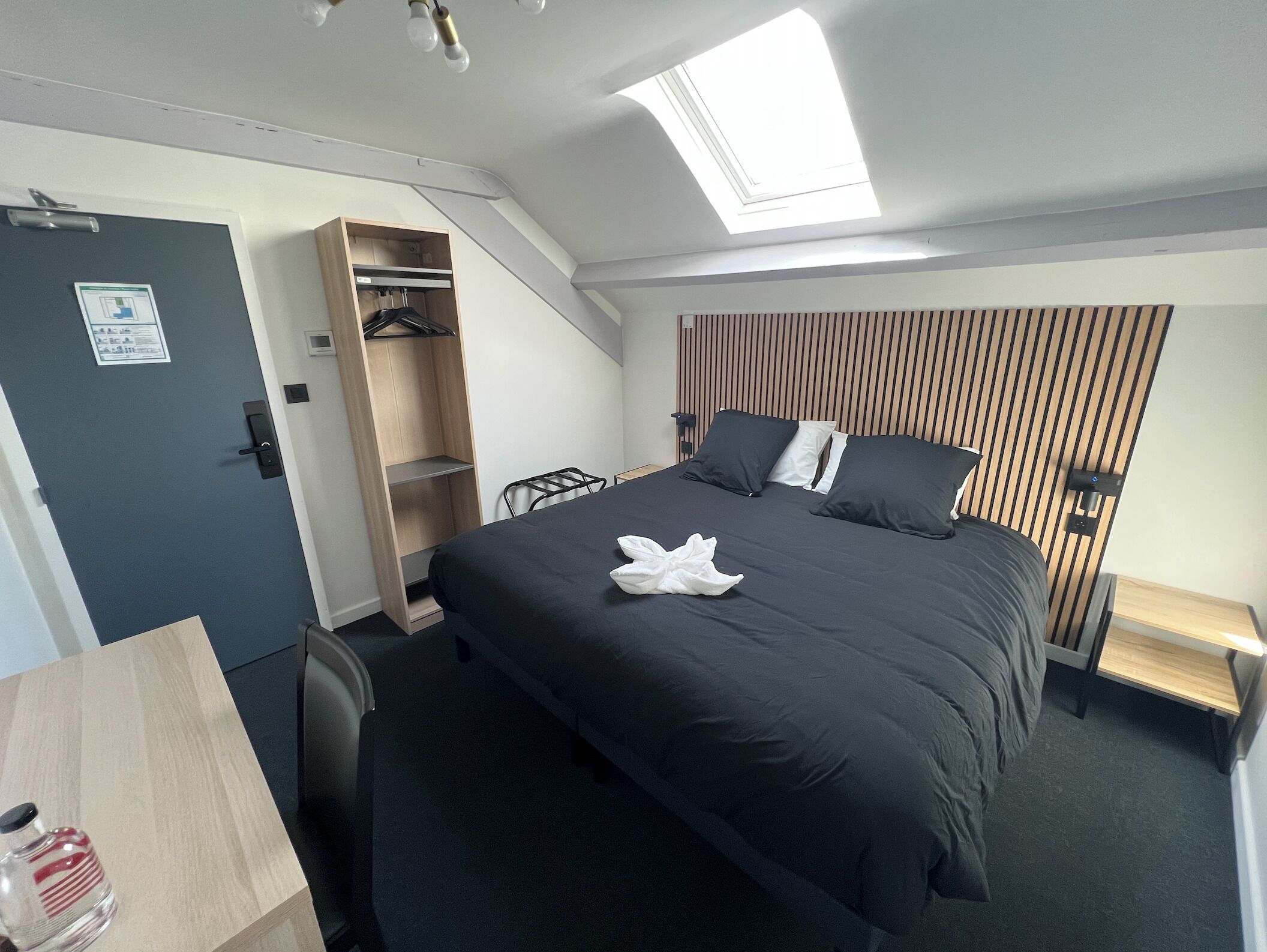 A compact hotel room with a bed, a desk, a wardrobe, and a skylight.