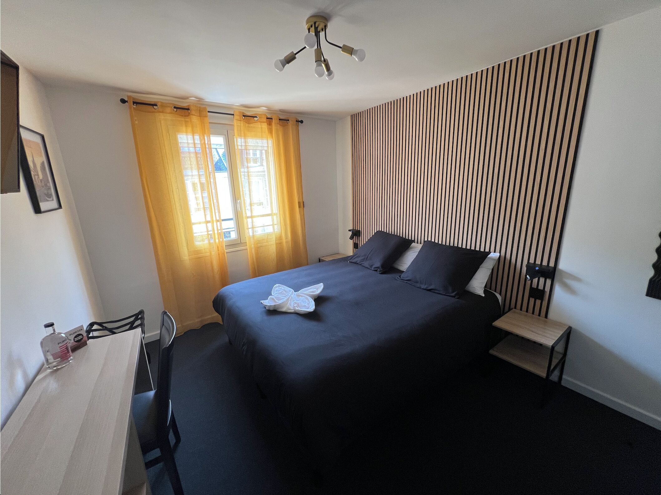 Habitación de hotel con cama, mesita de noche, silla, escritorio, lámpara de escritorio, ventilador de techo y ventana con cortinas.